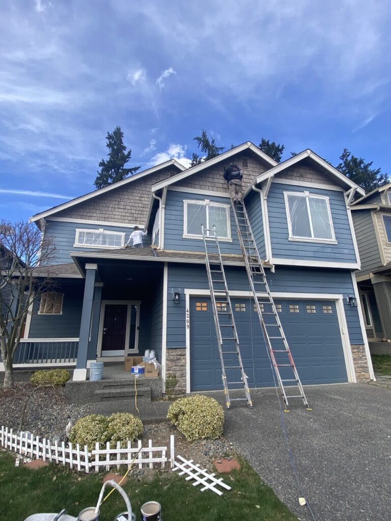 A person stands on a ladder painting the upper exterior of a two-story blue house with white trim. Various ladders and painting supplies are scattered on the driveway and front yard. The sky is partly cloudy.