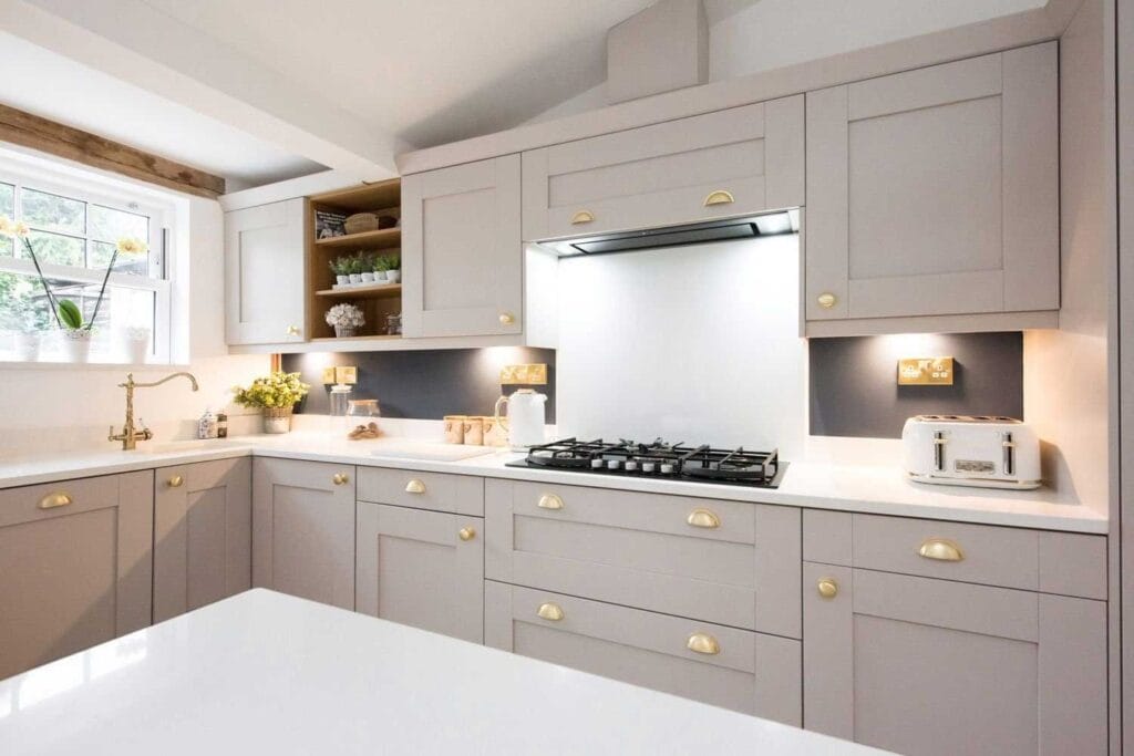 A modern kitchen with light gray cabinets, gold handles, a gas stove, white countertops, a white toaster, and a window with shelves holding plants and dishes. The space is bright and tidy.