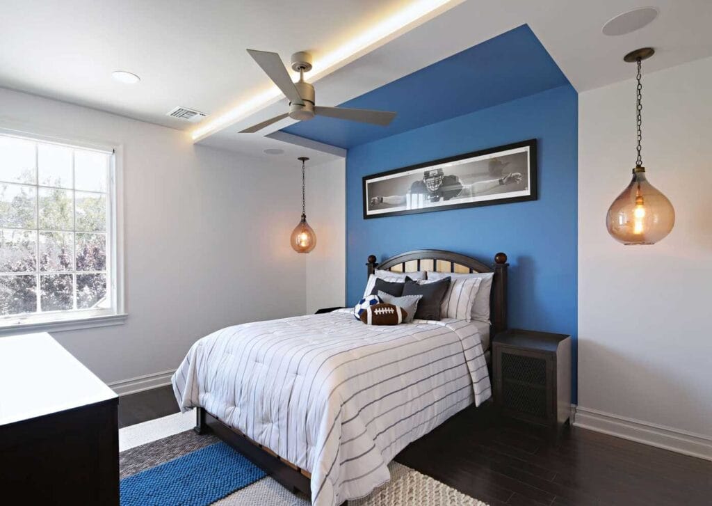 A modern bedroom with a blue accent wall behind a bed, striped bedding, a football pillow, two hanging pendant lights, a ceiling fan, a framed photo above the bed, and natural light from a large window.