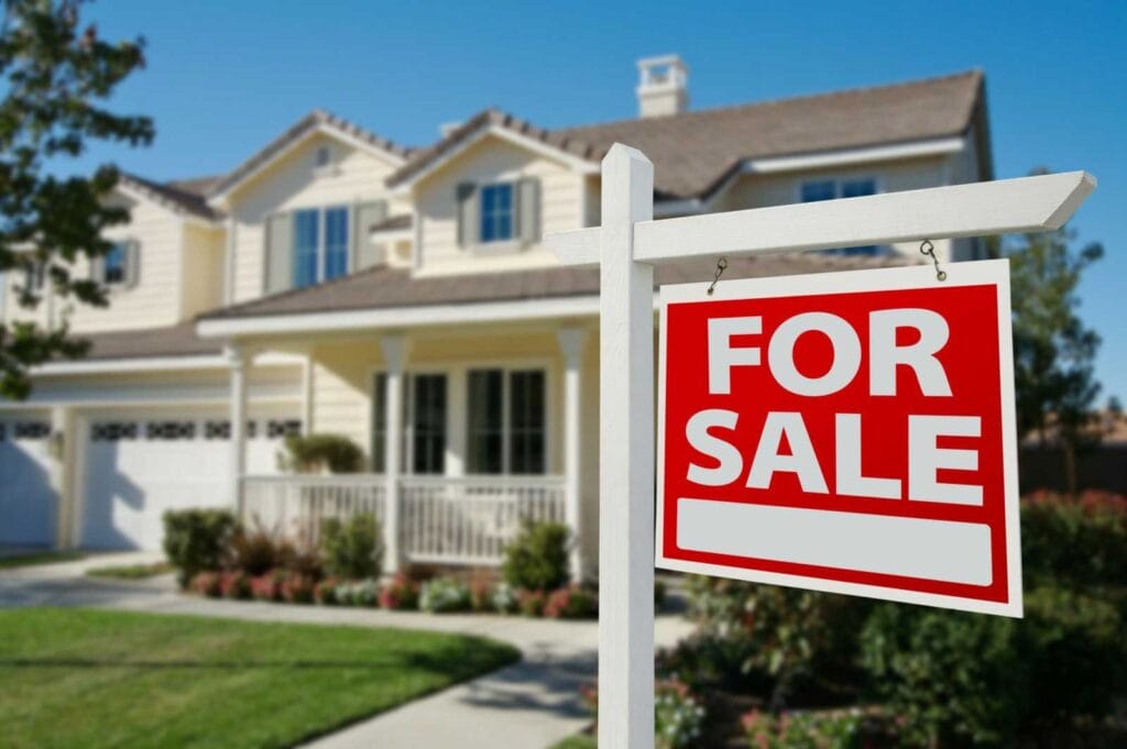 A red and white For Sale sign is displayed in front of a two-story suburban house with a porch, manicured lawn, and driveway on a sunny day.