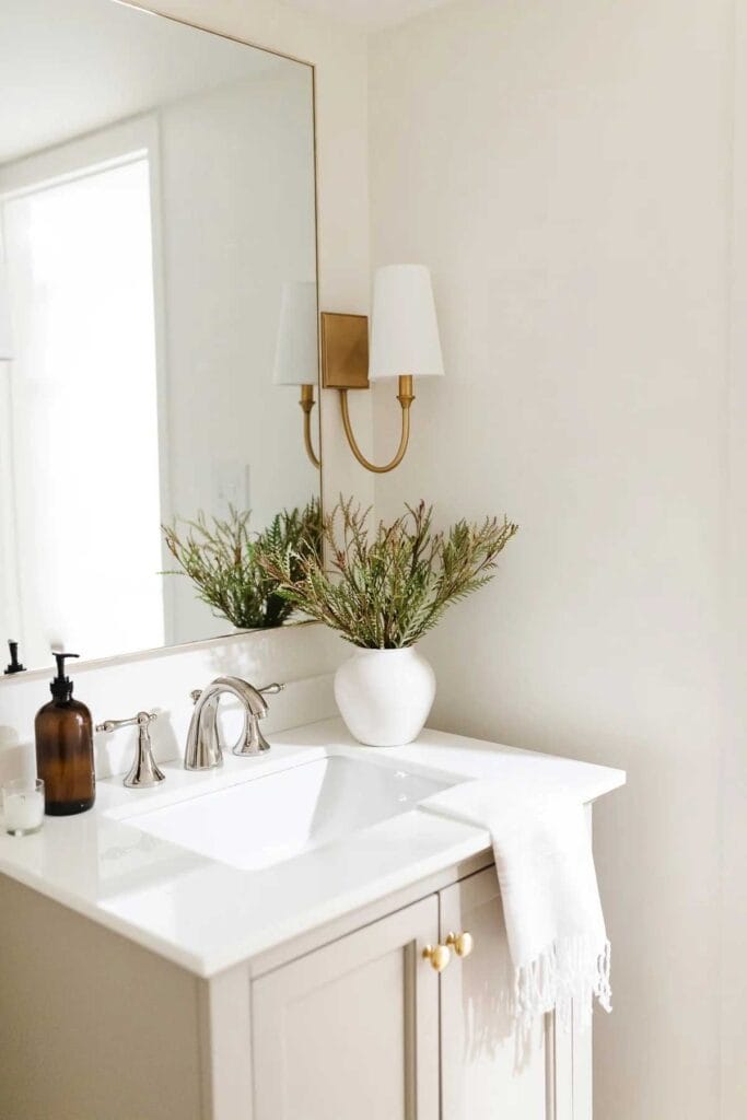 A bright, modern bathroom with a beige vanity, white countertop, chrome faucet, and a large mirror. A white towel hangs on the counter, with a white vase of greenery and a soap dispenser beside the sink.