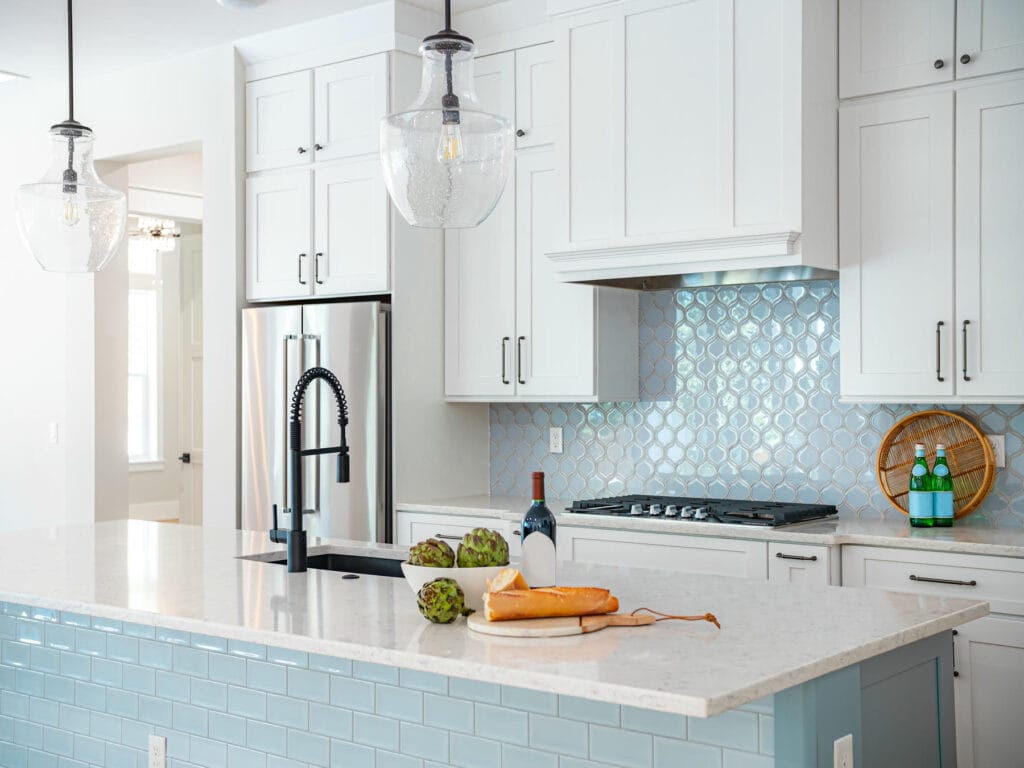 Modern kitchen with white cabinets, stainless steel appliances, blue tiled backsplash and island. On the island are a baguette, artichokes, a bottle of wine, and a basket with bottled drinks. Two pendant lights hang above.