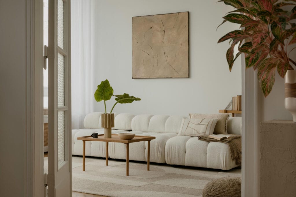 A modern living room with a white, plush sofa, a wooden coffee table holding a vase with large green leaves, neutral decor, a beige abstract painting, and soft natural light streaming through sheer curtains.