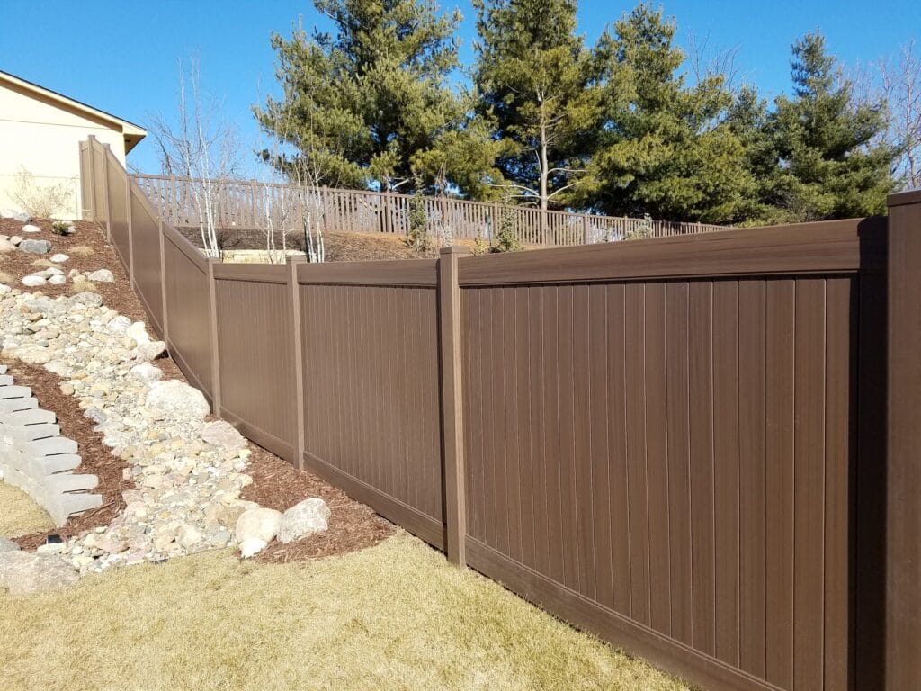A tall, brown privacy fence runs along a sloped backyard with rocks and mulch on one side, and pine trees and blue sky in the background.