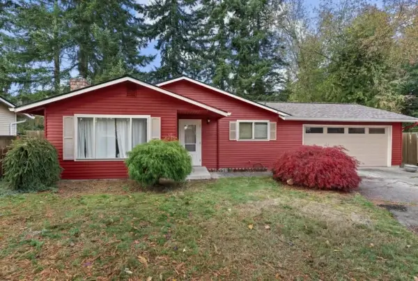 A single-story red house with white trim, large front window, and attached garage. The yard has green grass, a small tree, and red and green bushes. Tall evergreen trees and neighboring houses are in the background.