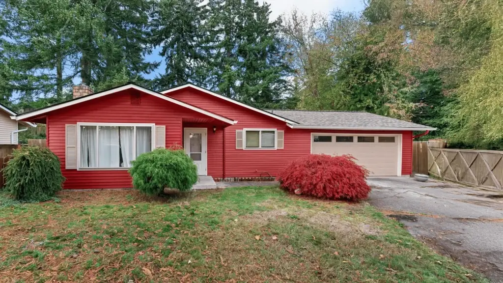 A small red house with white trim, a double garage, and large front windows sits amid green and red bushes, with trees and a wooden fence in the background.