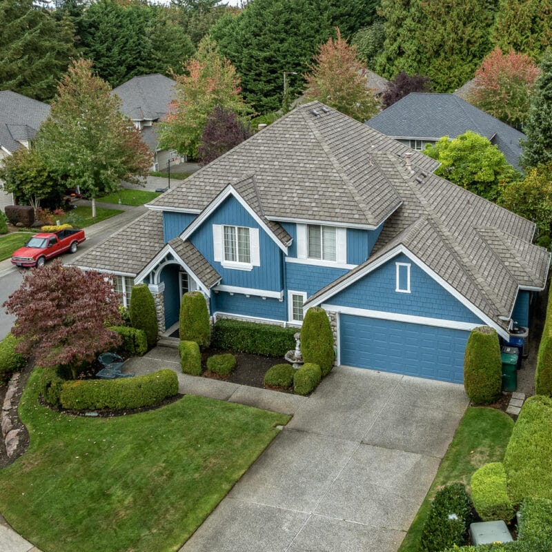 Aerial view of a suburban neighborhood featuring a blue two-story house with a well-manicured lawn, shrubs, and a curved driveway, surrounded by other houses and green trees.