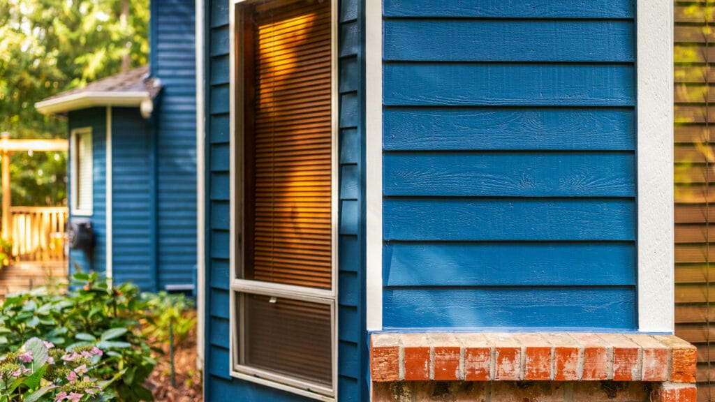 Close-up of the exterior of a house with blue wooden siding, a brick windowsill, and windows with brown blinds. Green plants are visible in the foreground and a wooden deck is in the background.