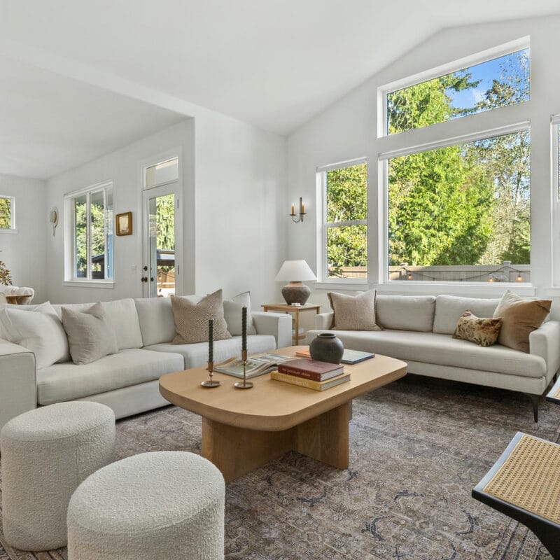 A bright, airy living room with white walls, large windows, neutral-toned furniture, a wooden coffee table, and minimalist decor. Light streams in, highlighting the cozy, modern space with outdoor greenery visible through the windows.