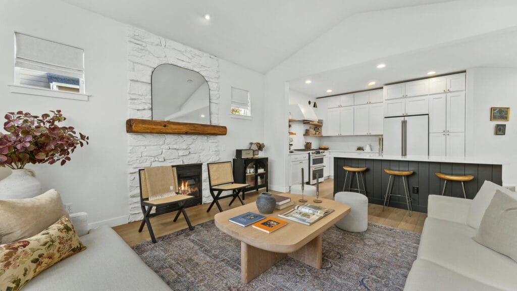 Modern living room with white walls, stone fireplace, wood mantel, a large mirror, cozy sofas, a light wood coffee table, two woven chairs, and an open kitchen with white cabinets and barstools in the background.