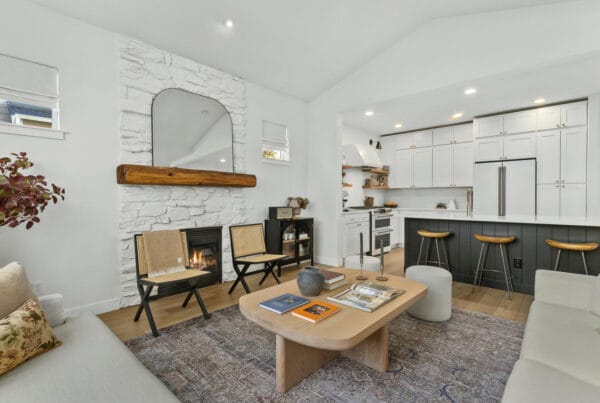 Modern living room with white walls, stone fireplace, wood mantel, a large mirror, cozy sofas, a light wood coffee table, two woven chairs, and an open kitchen with white cabinets and barstools in the background.
