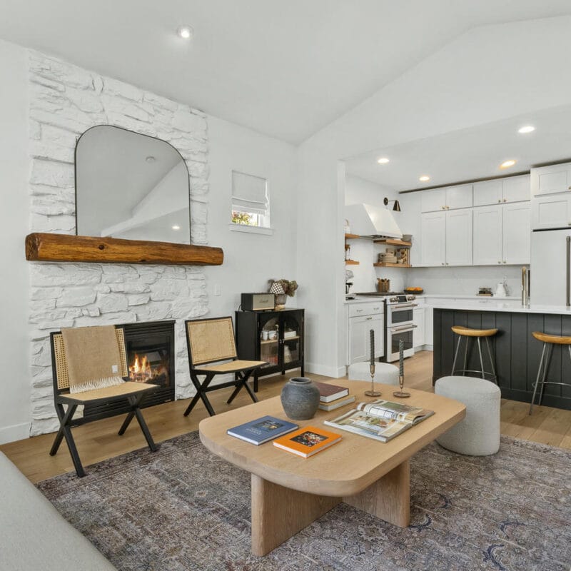 Modern living room with white walls, stone fireplace, wood mantel, a large mirror, cozy sofas, a light wood coffee table, two woven chairs, and an open kitchen with white cabinets and barstools in the background.