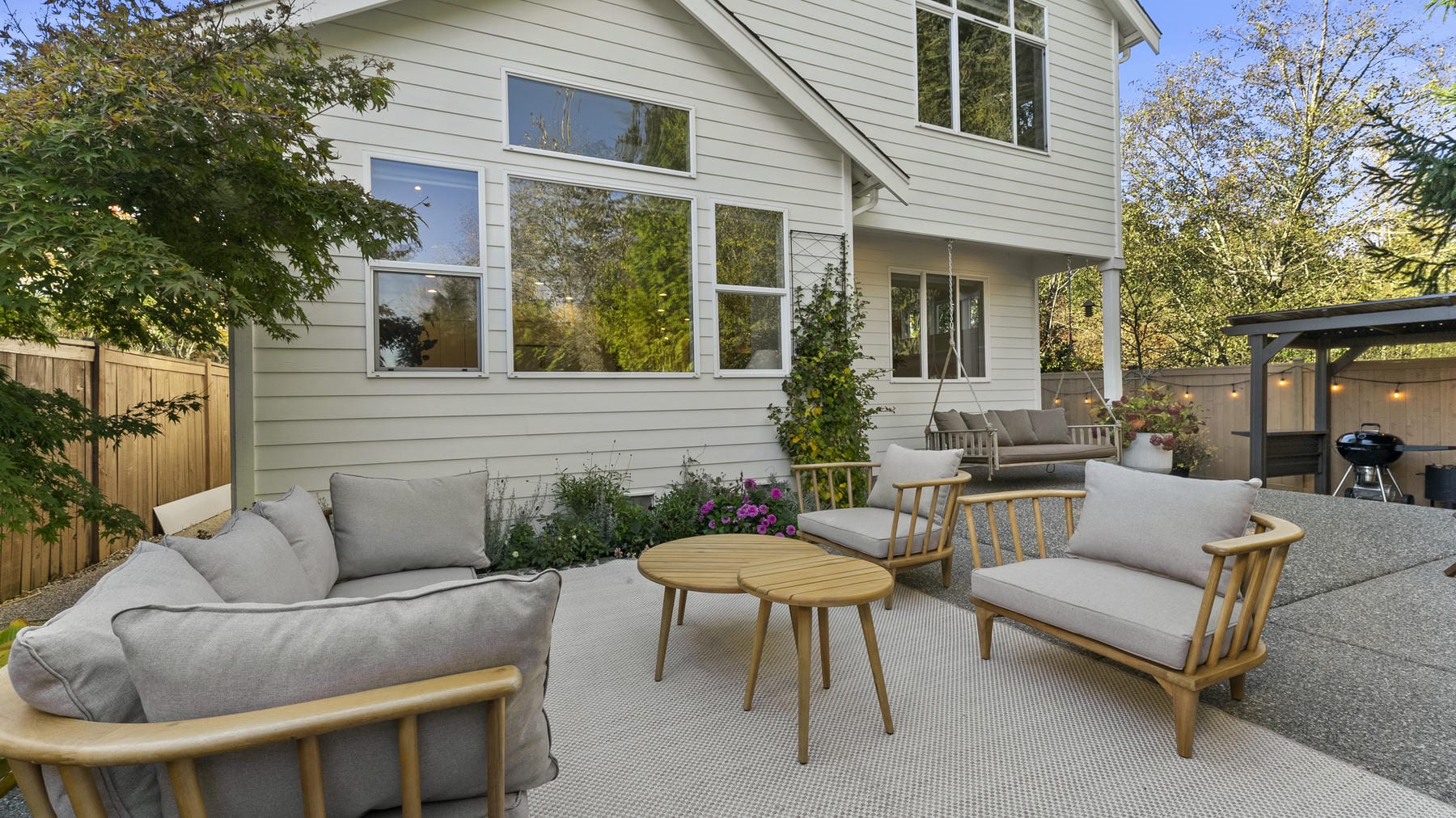 A modern backyard patio features light wood outdoor furniture with beige cushions, a round coffee table, and a neutral rug. The patio is surrounded by greenery and borders a white house with large windows.
