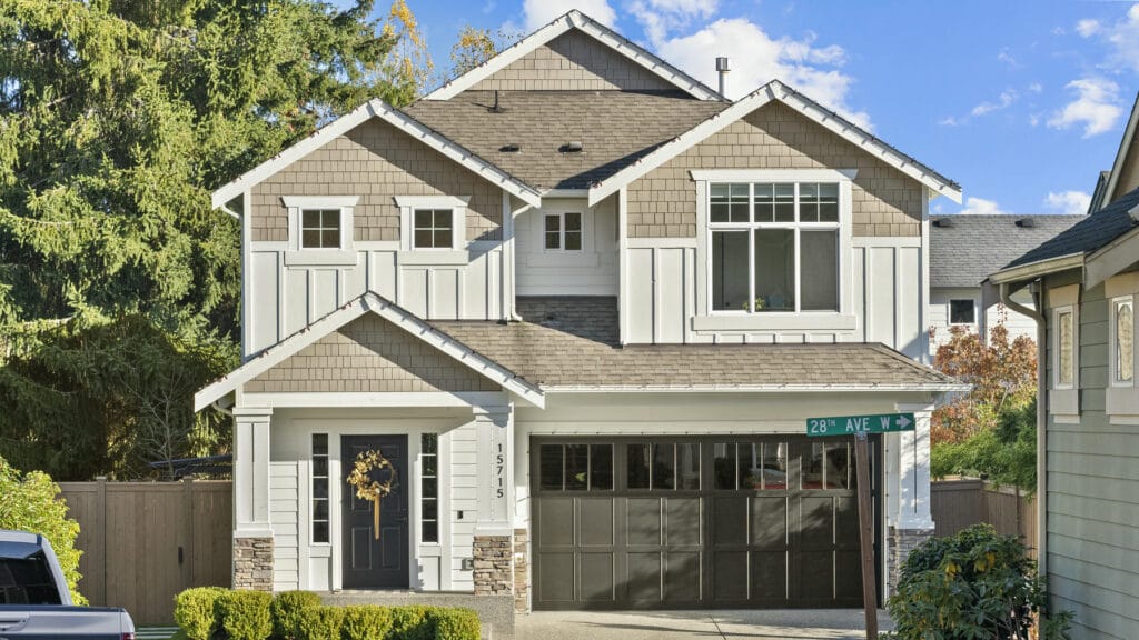 A two-story suburban house with gray siding, white trim, and stone accents. The front door has a wreath, and there’s a large garage. The house sits on a tidy lot with bushes and trees, at the corner of 28th Ave.