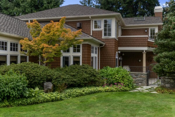 A large two-story house with brown shingle siding, white trim, and multiple windows, surrounded by lush green landscaping, shrubs, trees, and a well-maintained lawn.