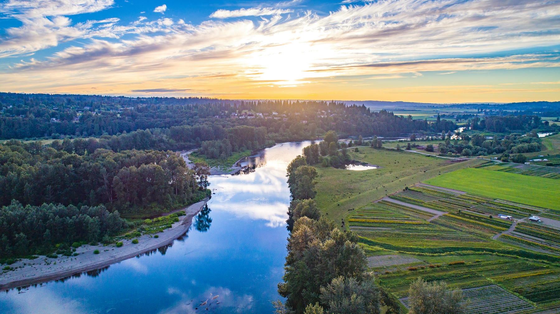 A scenic aerial view of a river winding through green fields and forests at sunset, with a partly cloudy sky and the sun low on the horizon, reflecting off the water.