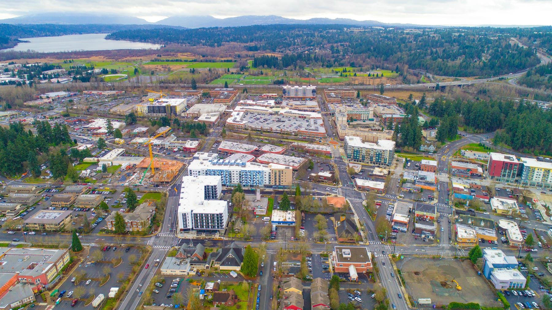 Aerial view of a suburban city with residential and commercial buildings, roads, and green spaces, surrounded by trees and hills, with a body of water visible in the background under a cloudy sky.