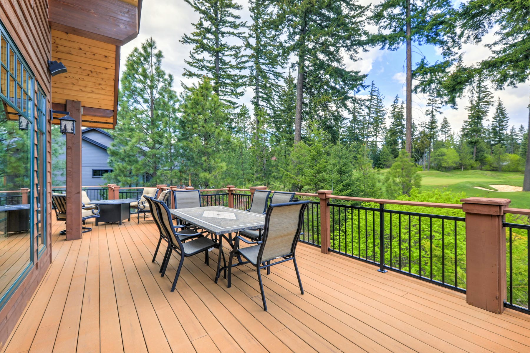 Spacious wooden deck with a dining table and chairs, surrounded by a black railing, overlooking a lush forest and golf course under a partly cloudy sky.