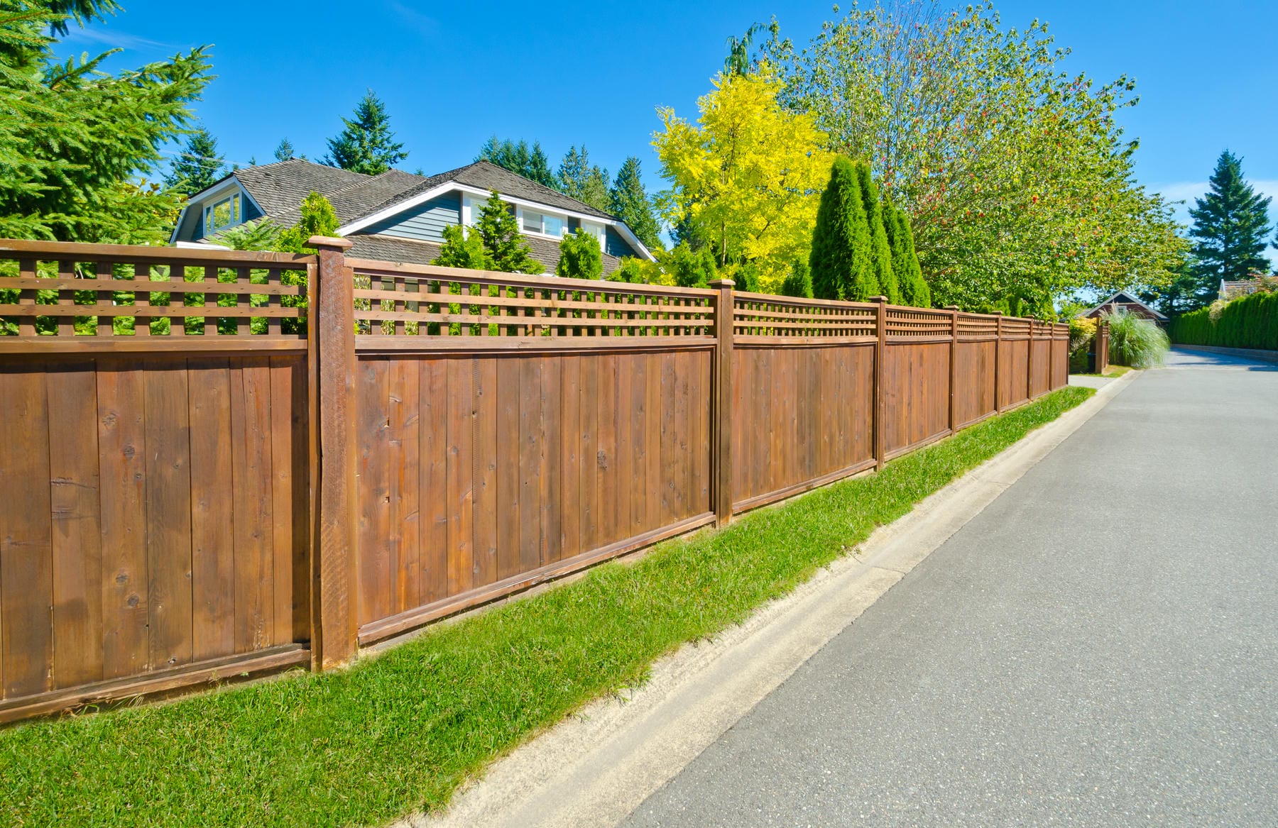 A wooden privacy fence lines the edge of a suburban street, enclosing a house and yard with green grass, trees, and shrubs under a clear blue sky.