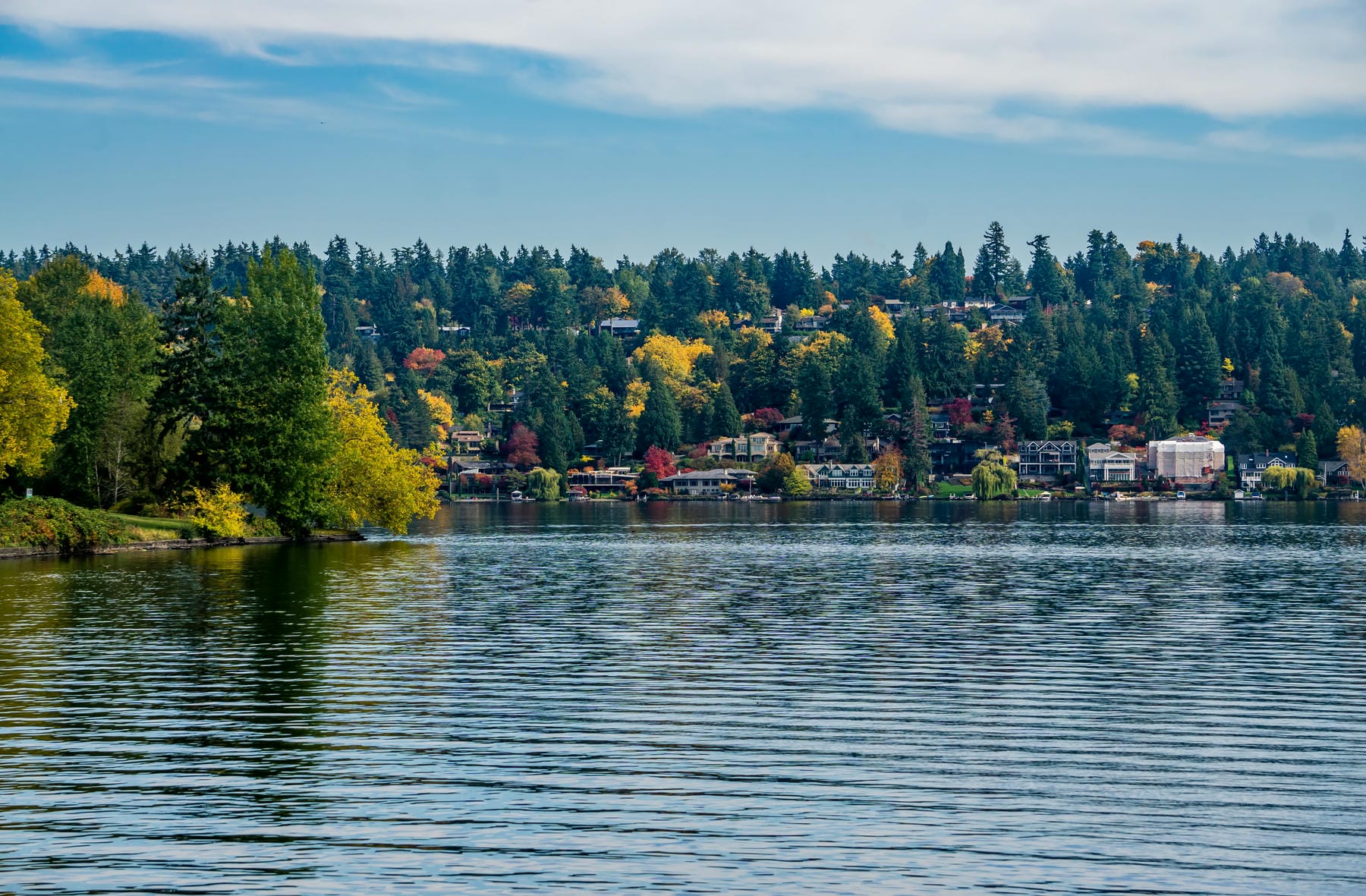A calm lake with gentle ripples reflects the trees with green and autumn colors lining the shore. Houses and buildings are nestled among the trees under a partly cloudy blue sky.