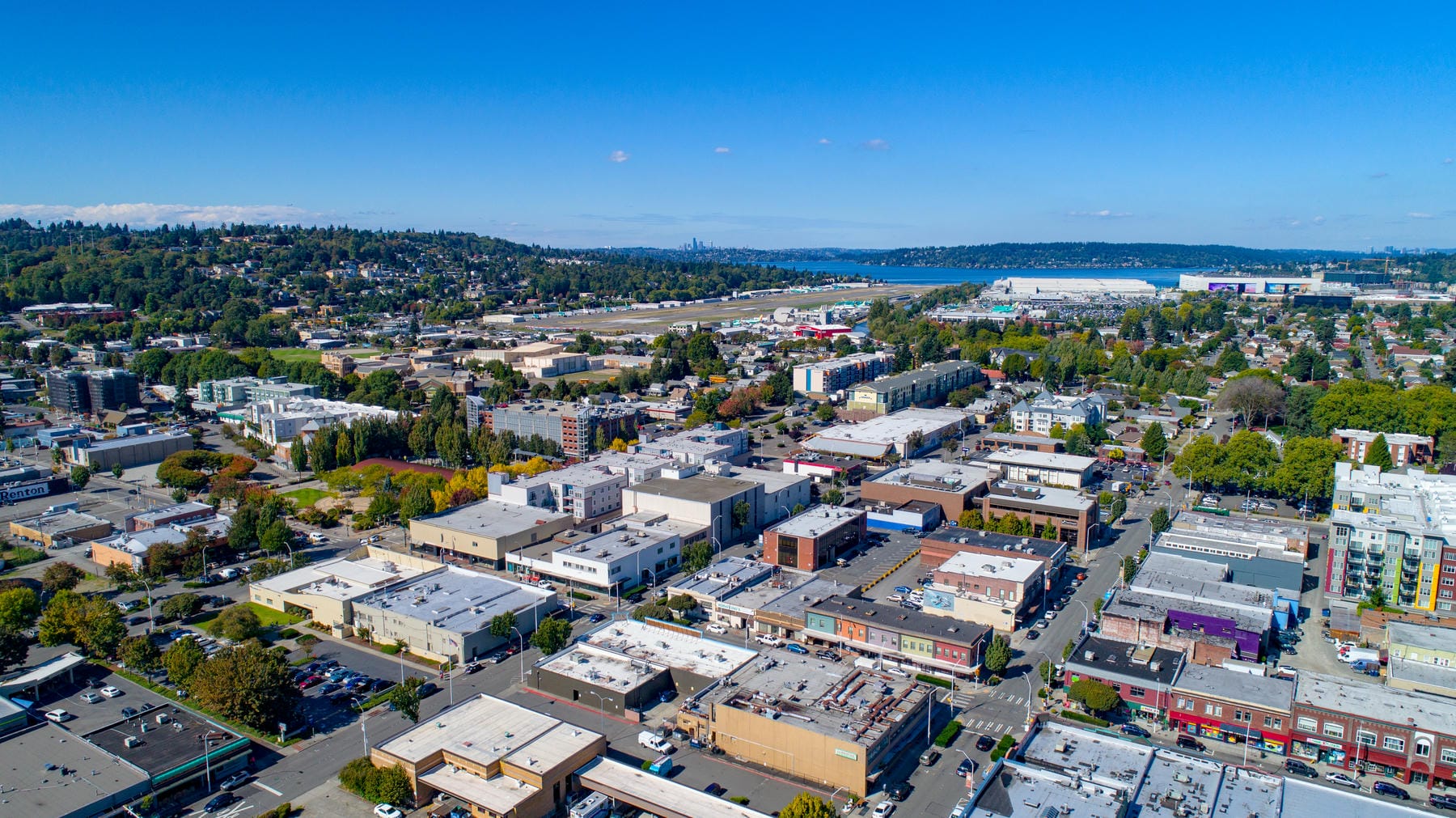 Aerial view of an urban neighborhood with commercial and residential buildings, tree-lined streets, and a distant waterfront under a clear blue sky. Hills and green areas can be seen in the background.
