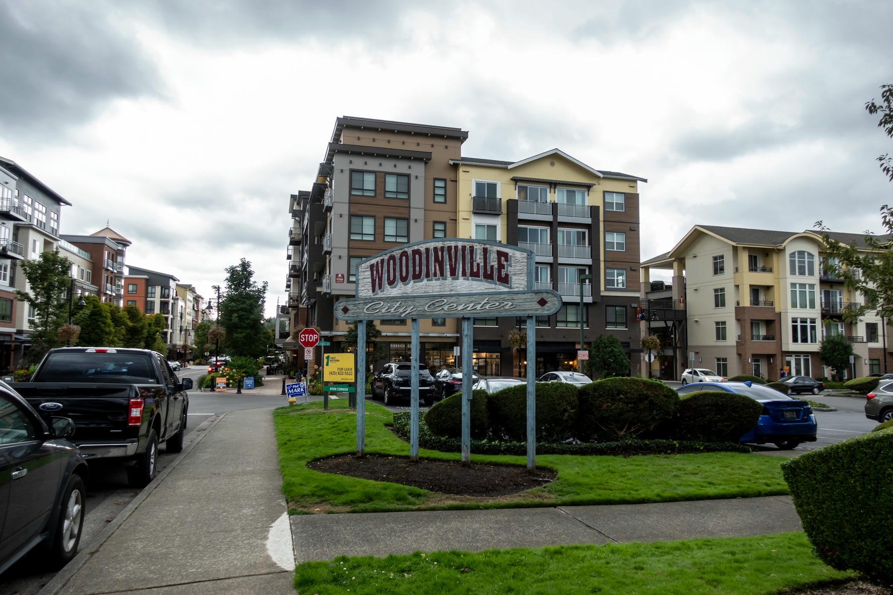 A large sign reading Woodinville City Center stands on a grassy median with modern apartment buildings, parked cars, and a cloudy sky in the background.