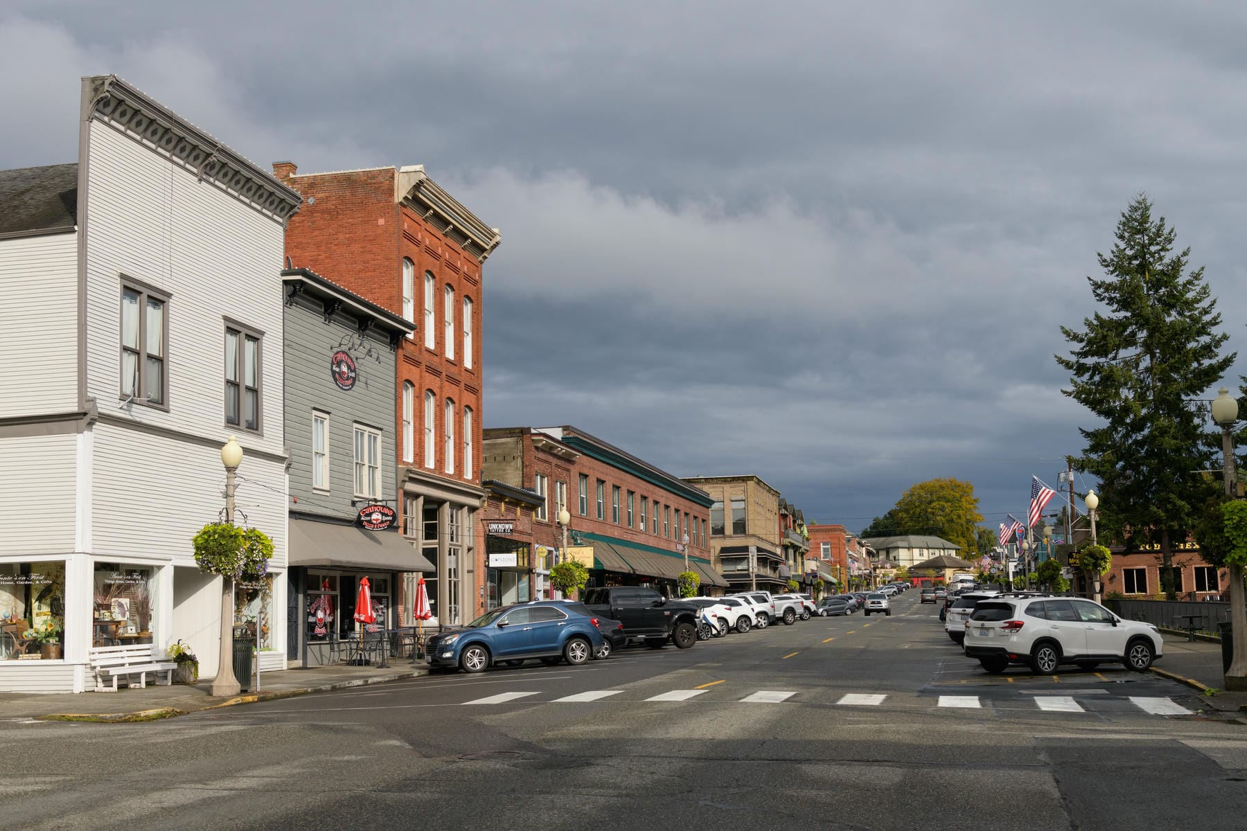 A small town main street with historic brick and wooden buildings, shops, parked cars, American flags, and a crosswalk under a cloudy sky. Trees with fall foliage are visible in the distance.