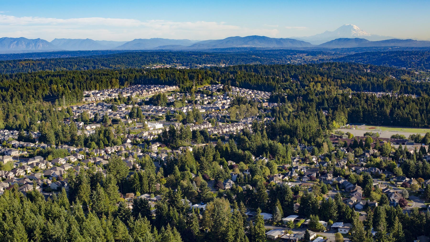 Aerial view of a suburban neighborhood surrounded by dense green trees, with mountains visible in the distance under a clear blue sky.
