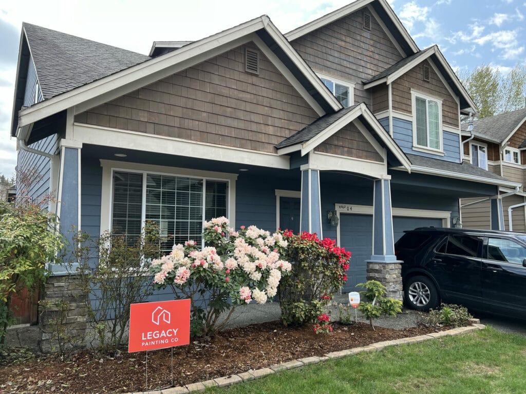 A two-story house with blue siding and brown shingles, white trim, and a front garden with blooming flowers. A black SUV is parked in the driveway, and a red Legacy Painting Co sign is placed in the yard.