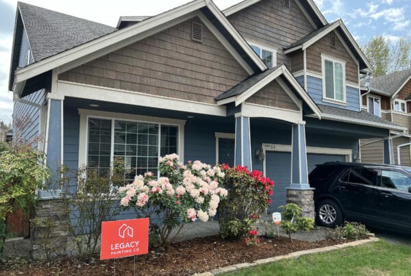 A two-story house with blue siding and brown shingles, white trim, and a front garden with blooming flowers. A black SUV is parked in the driveway, and a red Legacy Painting sign is placed in the yard.