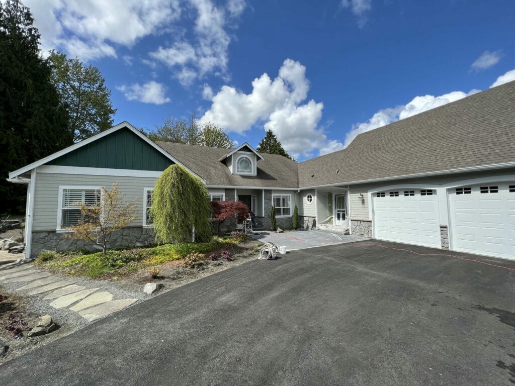 A modern suburban house with gray siding, green accents, white trim, and a large attached three-car garage. The driveway is paved, and landscaped garden beds with small trees border the entrance under a partly cloudy sky.