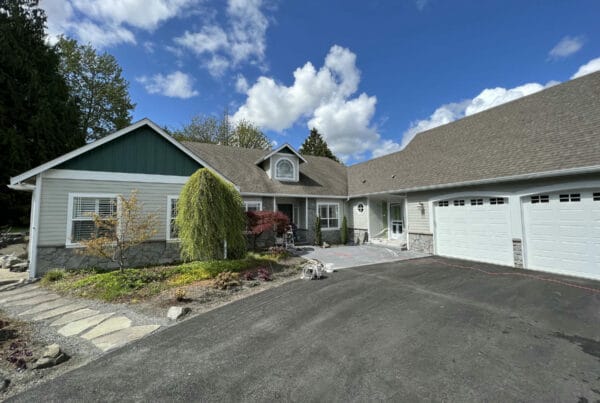 A modern suburban house with gray siding, green accents, white trim, and a large attached three-car garage. The driveway is paved, and landscaped garden beds with small trees border the entrance under a partly cloudy sky.