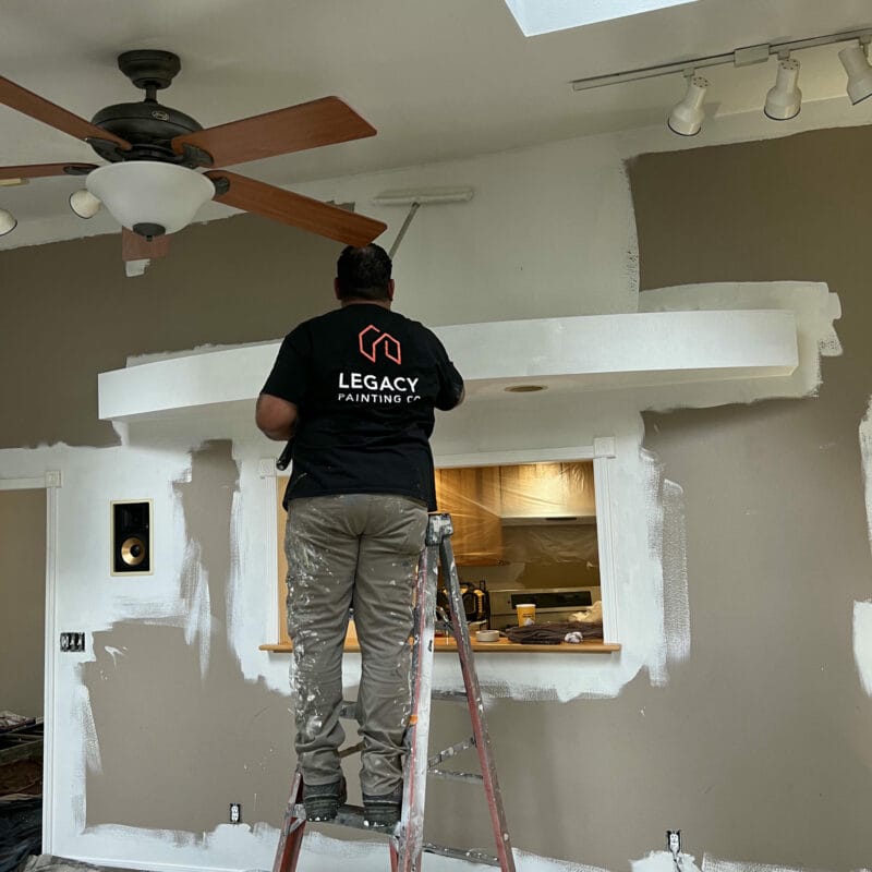 A painter, standing on a ladder, uses a roller to paint a brown wall white. He wears a black Legacy Painting shirt and paint-splattered pants. Painting supplies cover the floor, and the ceiling fan is visible above.