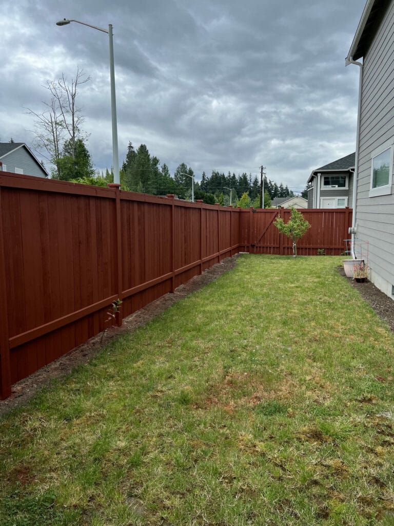 A backyard with a red wooden fence, green grass, a small tree, and a gray house on the right under a cloudy sky. Houses and trees are visible in the background.