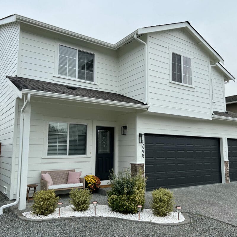 Two-story white house with black garage doors and a dark front door. There’s a small porch with a light pink bench, potted flowers, and neatly trimmed bushes along a gravel path leading to the entrance.
