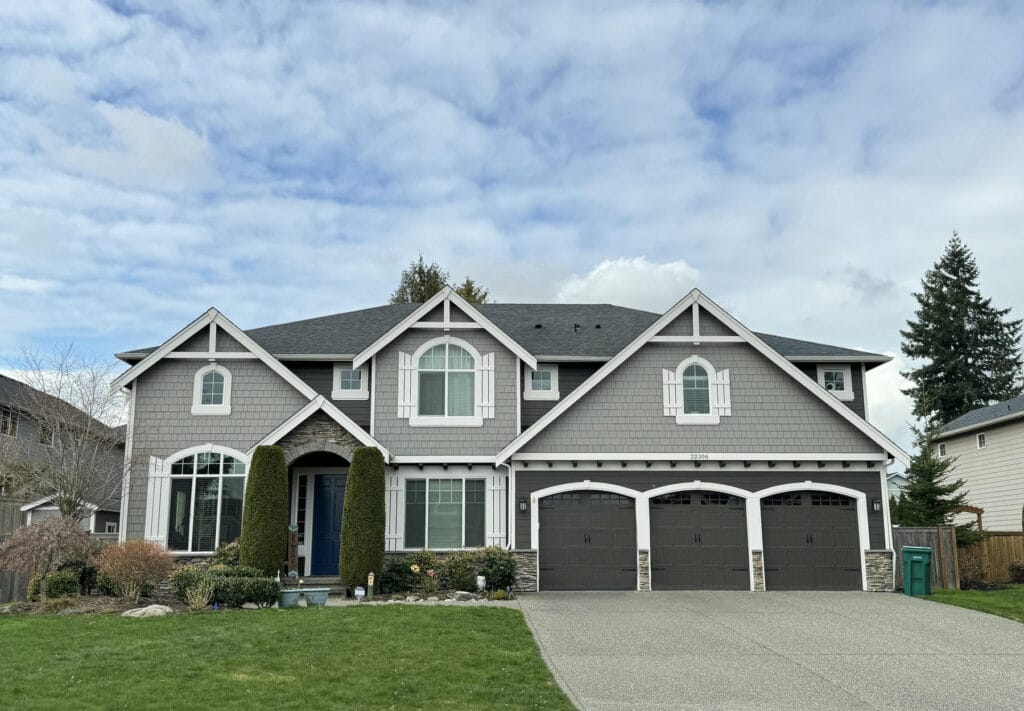 Two-story suburban house with gray siding, white trim, three-car garage, and large windows. The front yard has green grass, shrubs, and trees, with a wide driveway leading to the garage.