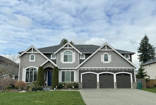 Two-story suburban house with gray siding, white trim, three-car garage, and large windows. The front yard has green grass, shrubs, and trees, with a wide driveway leading to the garage.