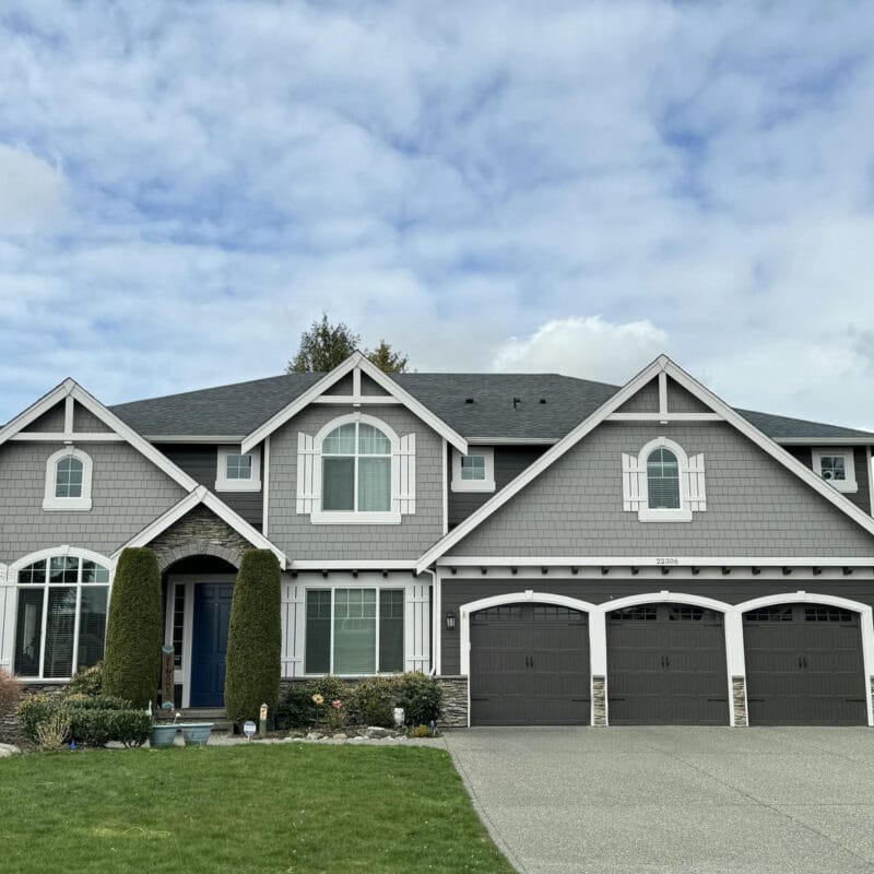 Two-story suburban house with gray siding, white trim, three-car garage, and large windows. The front yard has green grass, shrubs, and trees, with a wide driveway leading to the garage.