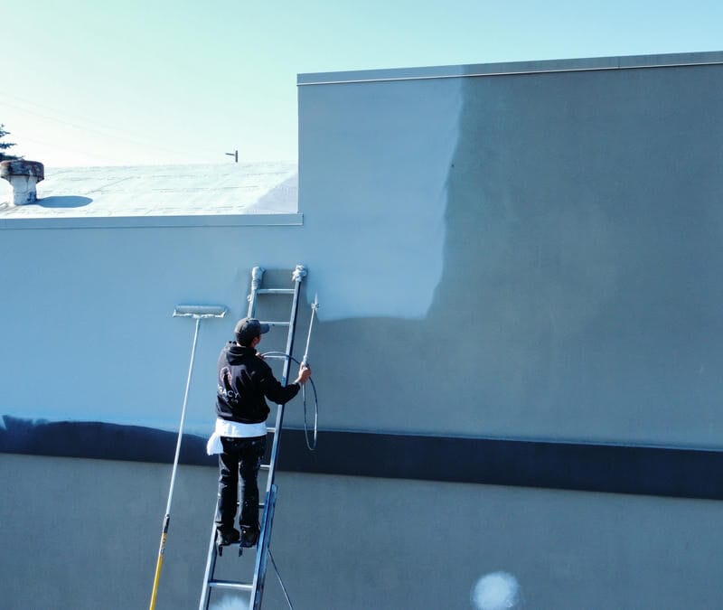 A person on a ladder uses a paint roller to paint a large exterior wall a light blue color, partially covering the original darker color. A for max sign is visible on the wall below.