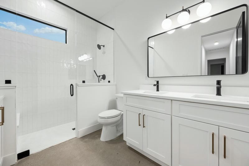 Modern bathroom with white cabinets, double sink, large rectangular mirror, glass-enclosed shower, white tiled walls, black fixtures, and a toilet under a frosted window. The space is bright and clean.