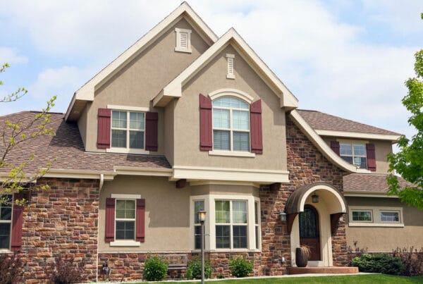 Two-story suburban house with tan stucco and stone exterior, brown shingle roof, white trim, and maroon window shutters. An arched entryway and manicured landscaping are visible in front.