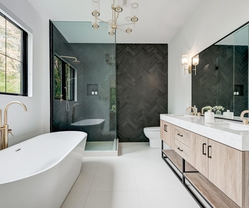 Modern bathroom with a freestanding bathtub, gold fixtures, a large double vanity with sinks, a glass-enclosed shower, a toilet, and large mirrors. Natural light comes through a window, illuminating the space.