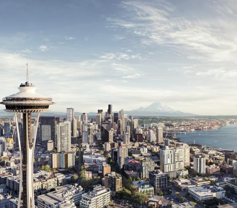 A panoramic view of downtown Seattle with the Space Needle in the foreground, skyscrapers, Elliott Bay, and Mount Rainier in the background under a partly cloudy sky.