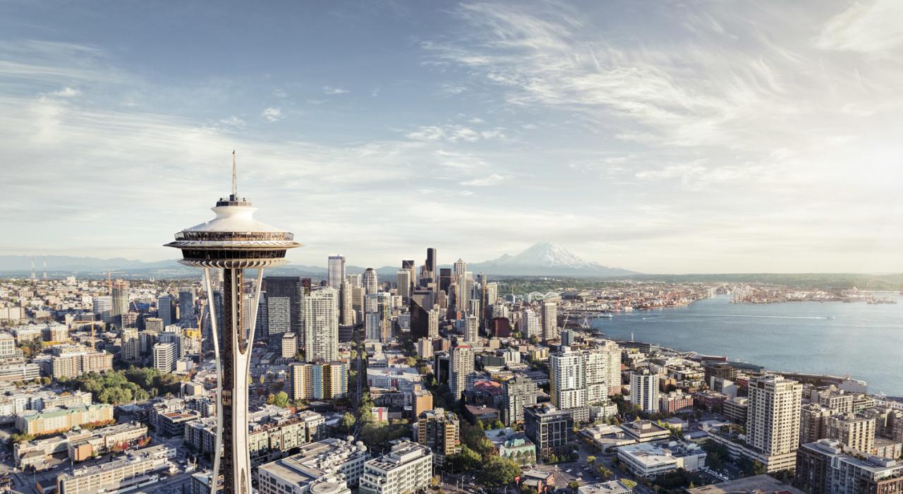 A panoramic view of downtown Seattle with the Space Needle in the foreground, skyscrapers, Elliott Bay, and Mount Rainier in the background under a partly cloudy sky.
