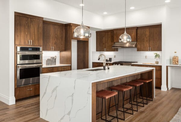 Modern kitchen with wood cabinets, stainless steel appliances, a large white marble island with a built-in sink, four brown stools, pendant lights, and hardwood floors.