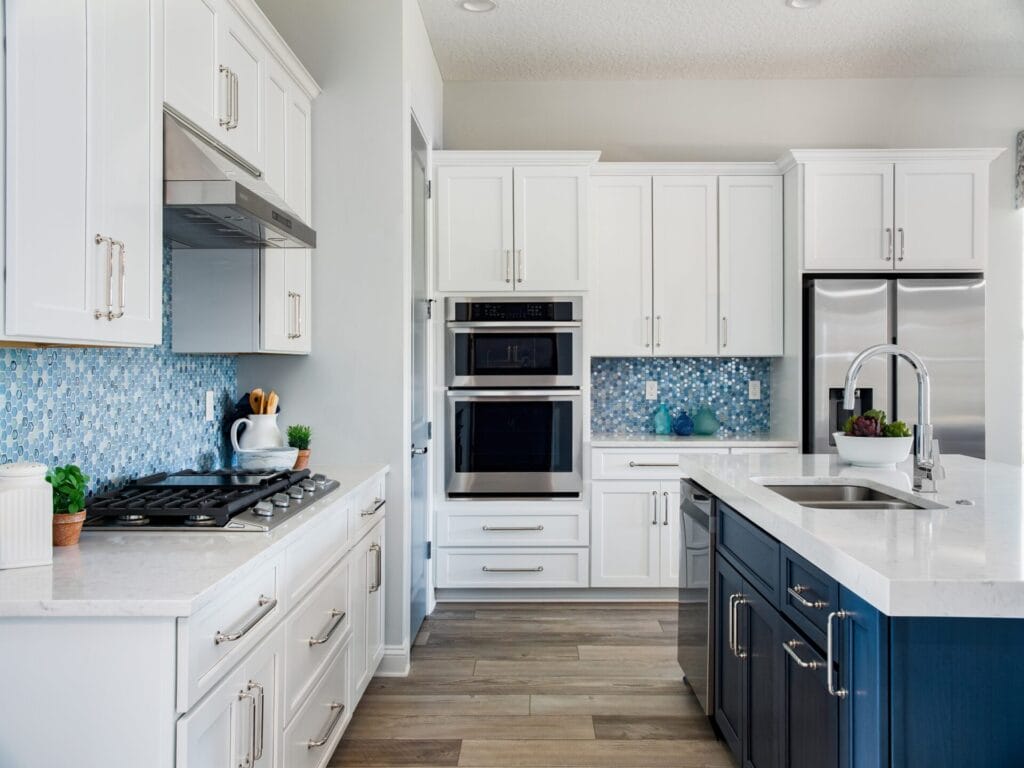 Modern kitchen with white cabinets, blue mosaic tile backsplash, stainless steel appliances, an island with a sink, and light wood flooring. Potted plants and kitchenware add decorative touches.