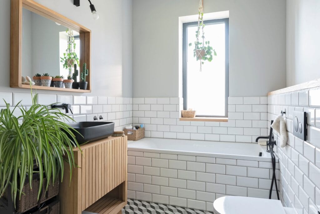 Modern bathroom with white subway tiles, a black sink on a wooden vanity, a rectangular mirror, potted plants, a bathtub under a window, and patterned floor tiles. Natural light brightens the space.