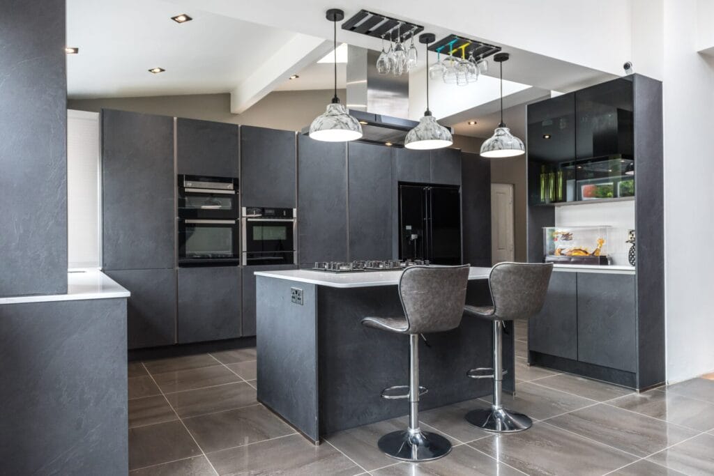 Modern kitchen with dark gray cabinets, a central island with two barstools, hanging pendant lights, built-in appliances, and a glossy tiled floor. Glass shelves with dishes and plants are visible in the background.