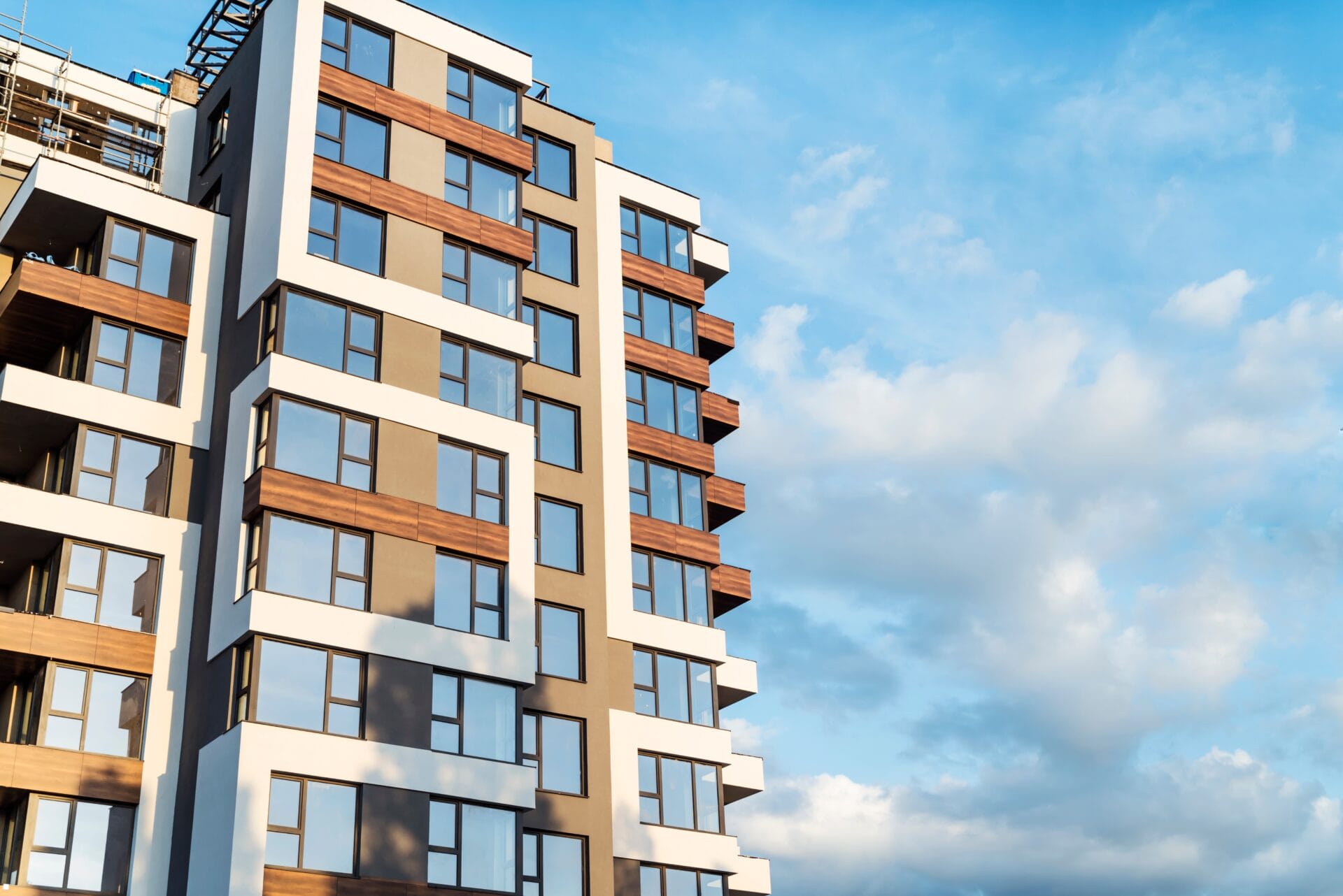 Modern multi-story apartment building with large windows and brown and white exterior accents, photographed against a blue sky with scattered clouds.