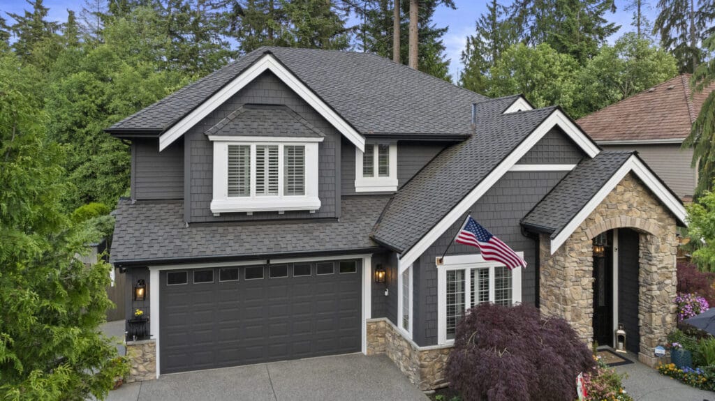 Two-story gray house with white trim, stone accents, and an American flag by the front entrance. Surrounded by trees and greenery, the house has a double garage and manicured landscaping.